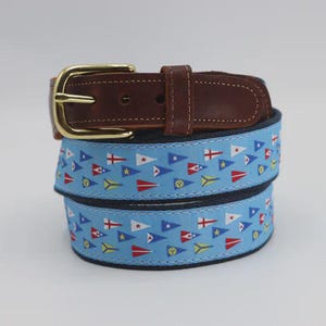 May include: A belt featuring a dark brown leather strap with a gold-toned metal buckle. The main body of the belt is light blue fabric decorated with a repeating pattern of colorful nautical signal flags in red, white, blue, and yellow.