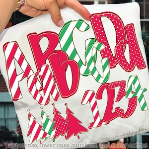 May include: A white fabric with a festive design featuring red and green letters, numbers, and Christmas trees. The letters and numbers have various patterns, including stripes and polka dots. The text "PNG ALPHABET SETS" is visible.