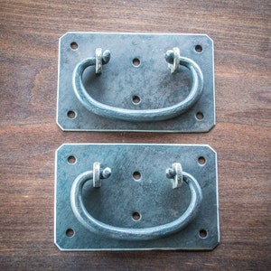 Puede incluir: Dos tiradores de puerta de metal plateado con una base rectangular redondeada y un tirador curvo. Los tiradores están montados en una superficie de madera.