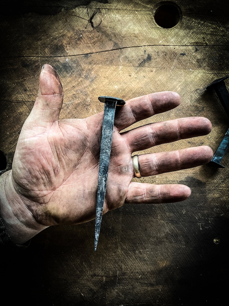 May include: A hand holding a large, black, hand-forged nail. The nail is about 15 cm long and has a square head. The nail is resting on a wooden surface.