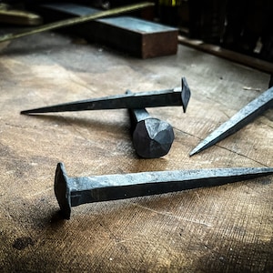 May include: Three hand-forged iron nails with different shaped heads, lying on a wooden surface.