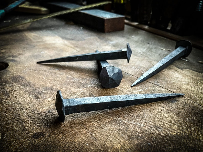 May include: Three hand-forged iron nails with different shaped heads, lying on a wooden surface.