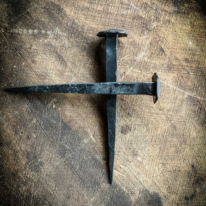 May include: Two black metal nails are crossed on a wooden surface. The nails are rusty and have a hammered finish.