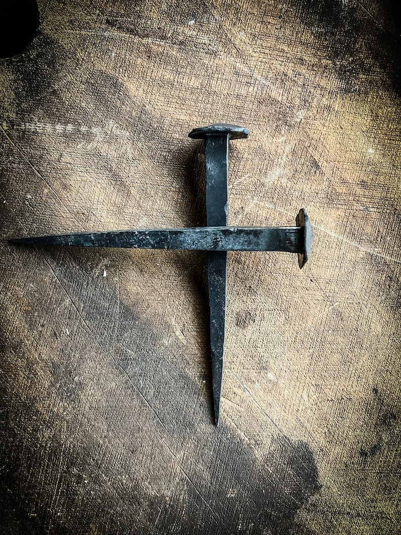 May include: Two black metal nails are crossed on a wooden surface. The nails are rusty and have a hammered finish.