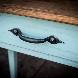 May include: Close-up of a wooden table with a light blue drawer. The drawer features a dark, ornate metal handle. The table has a natural wood top, creating a contrast with the painted drawer.