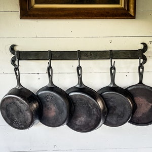 May include: Five cast iron skillets hang from a black metal rack mounted on a white wall. The skillets are all different sizes and have a rustic, well-used appearance.