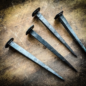 May include: Four antique iron nails with blunt points, arranged in a star shape on a wooden surface.