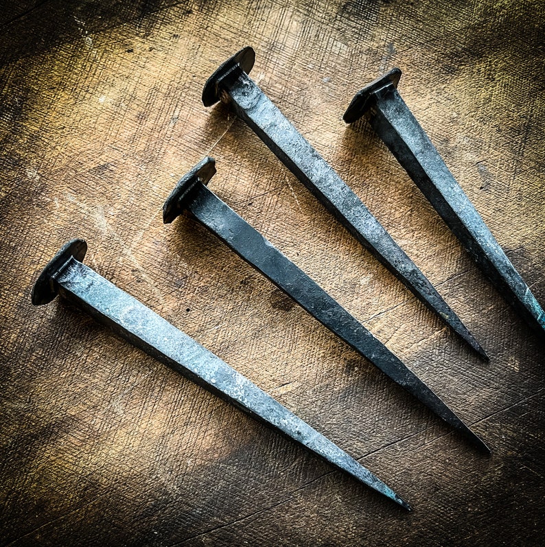 May include: Four antique iron nails with blunt points, arranged in a star shape on a wooden surface.