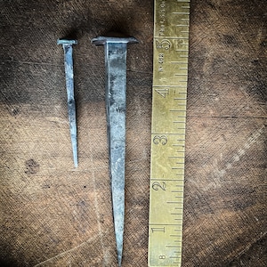 May include: Two metal nails of different sizes, one with a square head, the other with a pointed head, are placed on a wooden surface next to a brass ruler marked with inches. The ruler is labelled "The L.S. Co. No. 468".