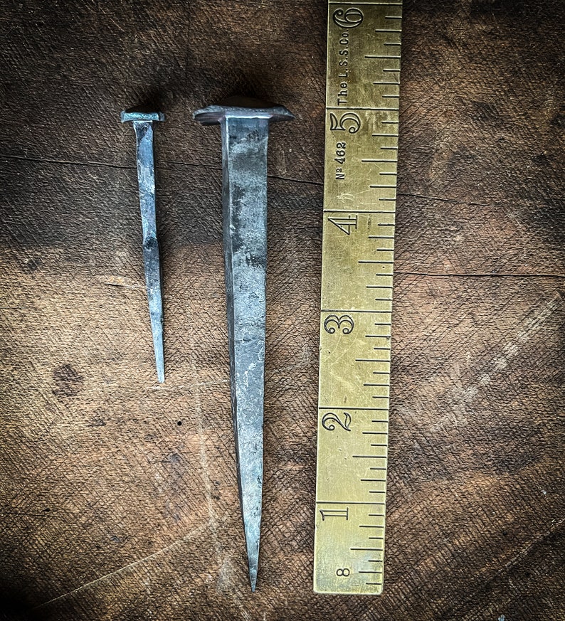 May include: Two metal nails of different sizes, one with a square head, the other with a pointed head, are placed on a wooden surface next to a brass ruler marked with inches. The ruler is labelled "The L.S. Co. No. 468".