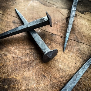 May include: Three antique iron nails with square heads, lying on a wooden surface. The nails are rusty and have a worn, aged appearance.