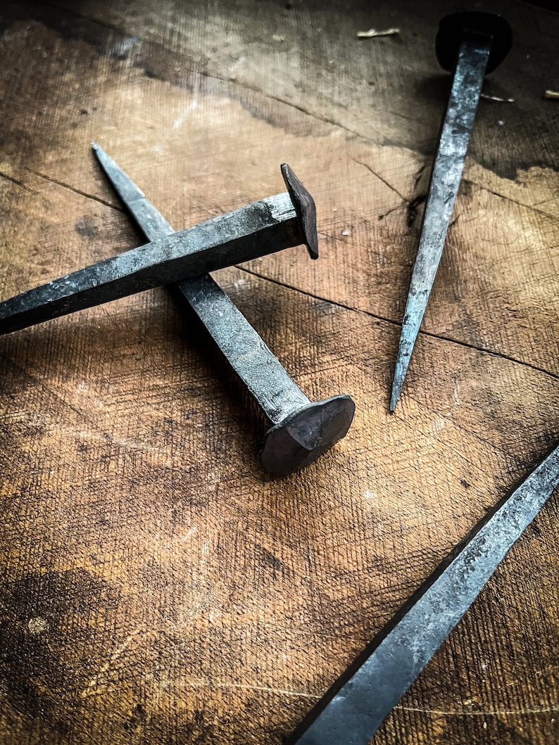 May include: Three antique iron nails with square heads, lying on a wooden surface. The nails are rusty and have a worn, aged appearance.