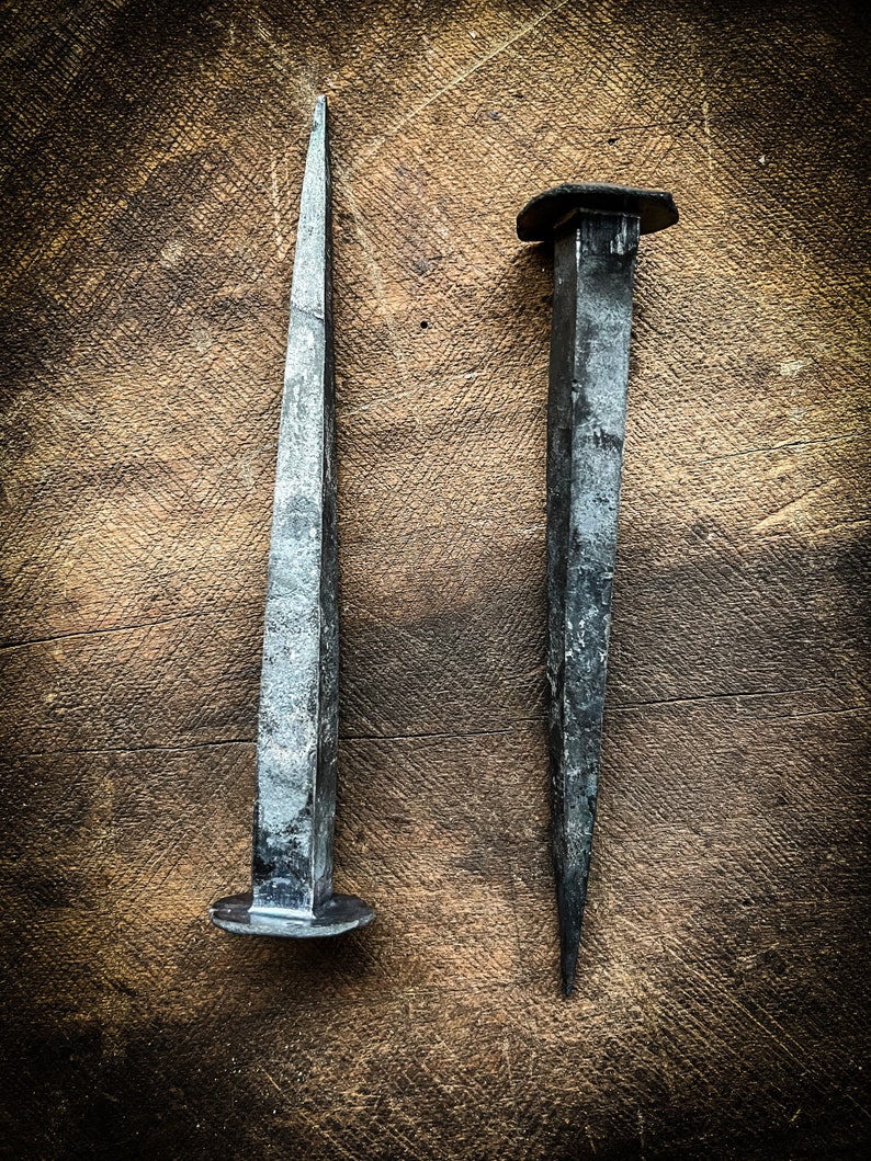 May include: Two antique metal nails with pointed tips and flat heads. The nails are on a brown wooden surface.