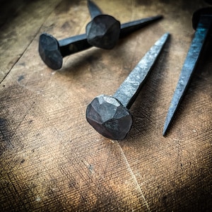May include: Three hand-forged iron nails with square heads, lying on a wooden surface. The nails are a dark grey colour and have a rustic, antique look.