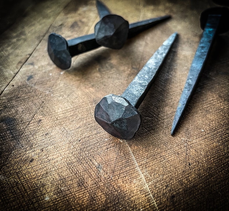 May include: Three hand-forged iron nails with square heads, lying on a wooden surface. The nails are a dark grey colour and have a rustic, antique look.
