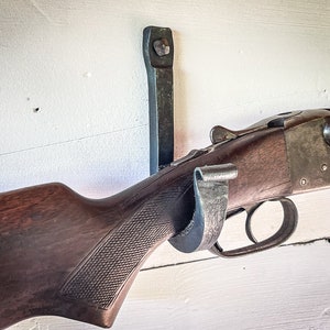 May include: A shotgun with a wooden stock and a metal barrel is mounted on a metal wall hook. The shotgun is resting on the hook with the barrel pointing to the right.
