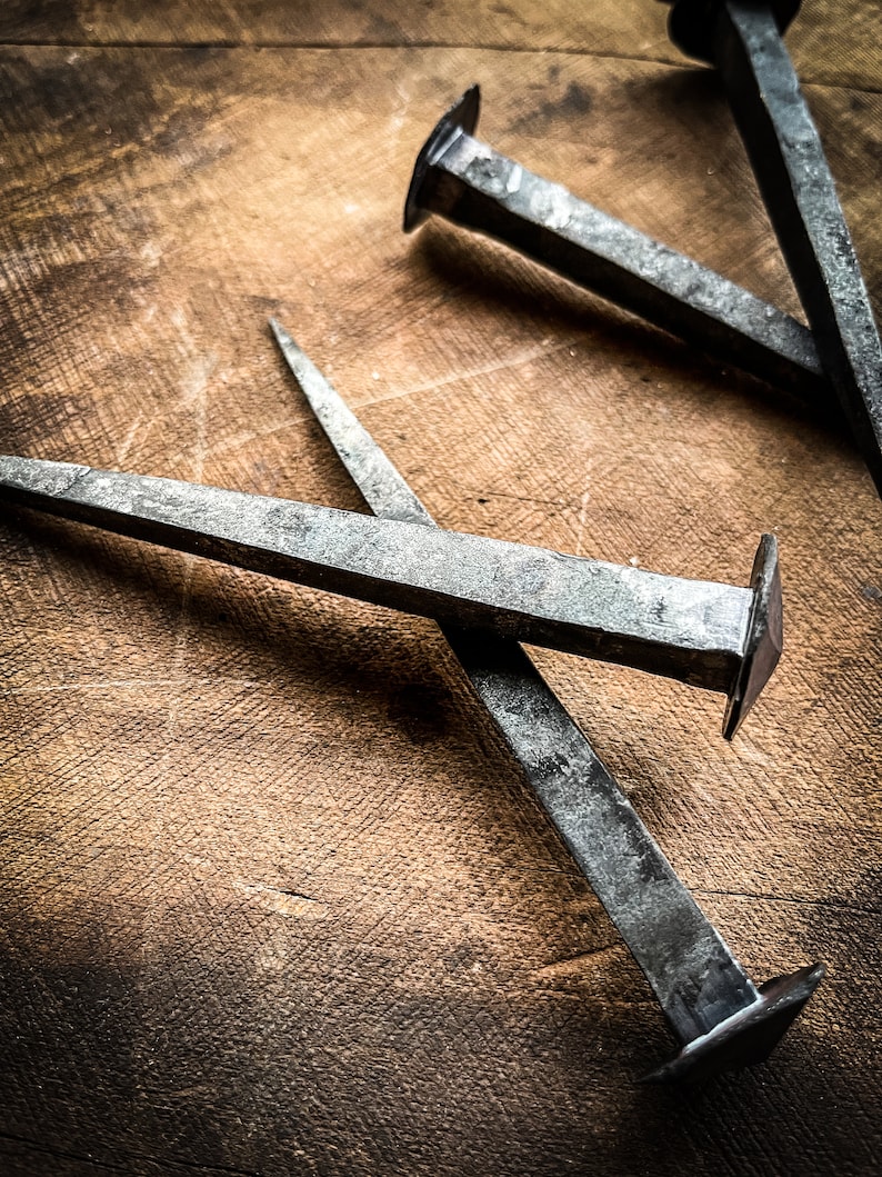 May include: Three antique-looking metal nails with square heads are arranged on a wooden surface. The nails are made of a dark metal and have a rustic appearance.