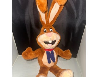 May include: A brown plush rabbit toy with long upright ears, a light tan muzzle, and a pink nose. The rabbit features a red collar with a large, dark blue letter N hanging from it. It has two visible white front teeth and a small pink tongue.