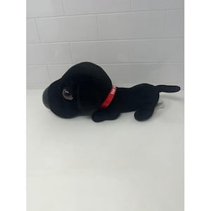 May include: A black plush dog toy with a red collar. The dog has large, round eyes and a small tail. The collar has white text. The toy is lying down on a white surface.