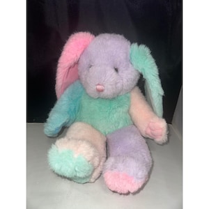 May include: A plush stuffed rabbit toy featuring a multi-coloured design with patches of pastel lavender, mint green, soft pink, and light blue fur. The rabbit has long floppy ears, a small pink stitched nose, and closed eyes with dark thread.