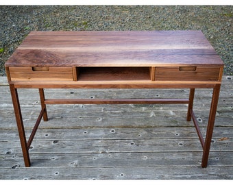 May include: A wooden desk with a natural finish. The desk has a rectangular top with two drawers and an open shelf. The legs are angled and the desk is made of a dark brown wood.