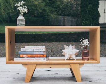 May include: A light-colored wooden coffee table with a rectangular design. The table features a lower shelf with books, a star-shaped decoration, and vases with flowers. The table is supported by angled legs.