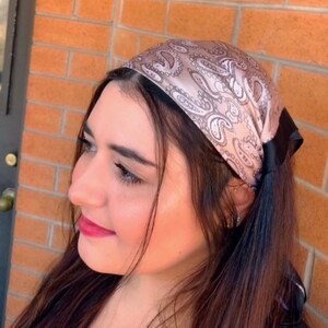 Puede incluir: Una diadema de color rosa con estampado de paisley y un lazo de satén negro. La diadema está puesta en la cabeza de una persona.