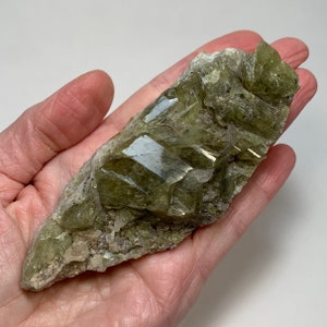 May include: A raw, uncut green crystal cluster with a crystalline structure. The mineral has a light green hue with reflective surfaces, and is set against a neutral background. The crystal is held in a hand.