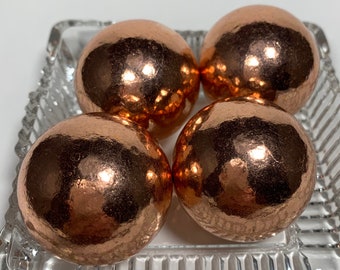 May include: Four copper-colored spheres with a textured surface are arranged in a clear, square, glass dish. The spheres have a metallic sheen, reflecting light. The dish has a decorative, ridged edge. The overall aesthetic is elegant and decorative.