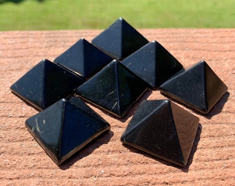 May include: Seven polished black obsidian pyramids arranged in a pyramid shape. The pyramids are square-based with smooth, reflective surfaces. The background is a blurred view of green foliage.