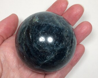 May include: A dark blue sphere with a polished surface, held in an open hand. The sphere has a marbled appearance with lighter blue and green inclusions. The object is a decorative item, possibly a gemstone or mineral specimen.