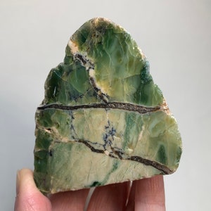 May include: A polished, irregularly shaped slab of green serpentine stone. The stone features a mix of light and dark green hues with black and brown veining. The surface has a natural, textured appearance, with visible cracks and variations in color.