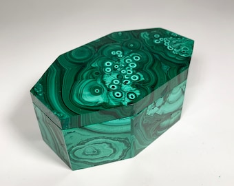 May include: A green malachite box with a geometric design. The box has a hinged lid and is made of polished stone. The surface displays a swirling pattern of dark and light green hues, creating a unique, natural aesthetic.
