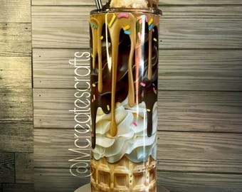 Könnte beinhalten: Ein Edelstahlbecher mit einem farbenfrohen Design eines Waffelhörnchen-Eisbechers mit Schokoladen- und Karamellsauce, Streuseln und Schlagsahne. Der Becher steht auf einem weißen Sockel mit verstreuten Schokoladenquadraten.