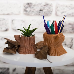 Könnte beinhalten: Zwei braune, aus Holz gefertigte Pflanzgefäße in Form eines Baumstumpfes. Ein Gefäß enthält eine kleine grüne Sukkulente. Das andere Gefäß enthält eine Gruppe bunter Stifte.