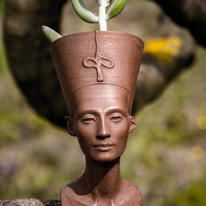 Op de afbeelding: Een bronzen plantenbak in de vorm van de buste van Nefertiti, met een vetplant die uit de bovenkant groeit. De plantenbak heeft een gedetailleerd gezicht en een hoge hoofdtooi. De achtergrond is wazig, met groene en bruine tinten.
