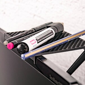 May include: A black plastic desk organizer with a honeycomb pattern is attached to the top of a black computer monitor. The organizer holds a black marker, a pink marker, and a pencil.