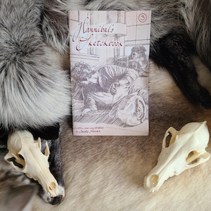 Może przedstawiać: Kopia "Hannibal's Sketchbook" z ilustracją w odcieniach sepii na okładce, przedstawiającą postać szkicującą. Dwie czaszki zwierząt i futrzane tło otaczają książkę. Książka jest oznaczona numerem "3".
