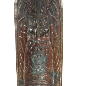 Puede incluir: Una pieza decorativa rectangular de bronce con un relieve detallado de una langosta. La langosta está posicionada centralmente, con sus pinzas y cola extendiéndose hasta los bordes. La pieza tiene una pátina oscura y envejecida.
