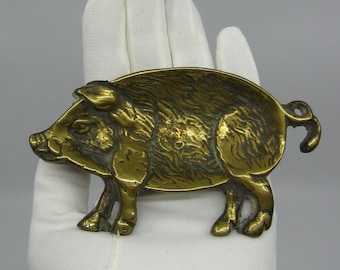 May include: A brass pig figurine with a textured surface. The pig is in a side profile, with its legs, snout, and curly tail visible. The figurine is held in a gloved hand against a gray background. The pig is approximately 6 inches long.