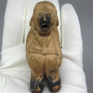 Könnte beinhalten: Eine kleine, braune Keramikfigur eines Menschen mit einem breiten Mund und schwarzen Augen. Die Figur trägt ein einfaches Outfit und schwarze Schuhe.