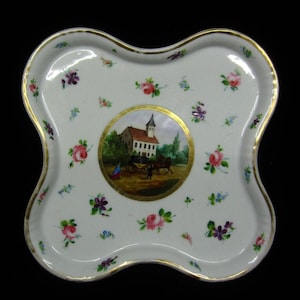 Puede incluir: Una bandeja de porcelana blanca con un borde dorado y un patrón floral. La bandeja tiene un medallón central con una escena pintada de un carruaje tirado por caballos frente a un edificio.