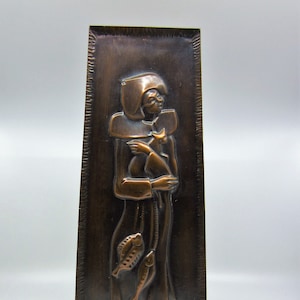 May include: A copper wall plaque with a raised relief design of a person holding a fish. The person is wearing a hooded robe and has a fish in each hand.