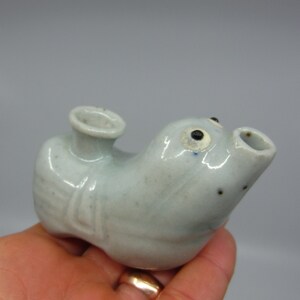 May include: A small, light blue ceramic figurine of a fish with black eyes. The figurine has a spout on the top and a hole on the bottom, suggesting it may be a pourer or a whistle.