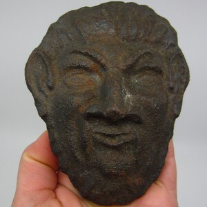 May include: A weathered, dark brown cast iron face sculpture with closed eyes and a slight smile. The sculpture has detailed facial features, including ears and textured skin. The piece is held in a hand against a plain background.