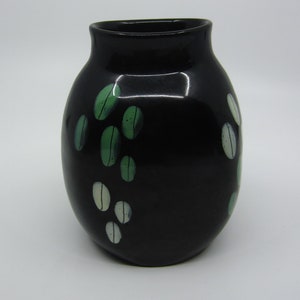 Puede incluir: Un jarrón de cerámica negro con forma redondeada y acabado brillante. El jarrón está decorado con hojas pintadas en verde y blanco.
