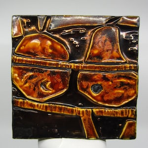 Puede incluir: Un azulejo de cerámica con una superficie texturizada. El azulejo es marrón y naranja con un fondo negro. El azulejo tiene un diseño geométrico.