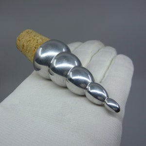 May include: A silver bottle stopper with a cork base. The stopper has a series of graduated, spherical shapes, creating an elongated design. The cork is light brown in colour.