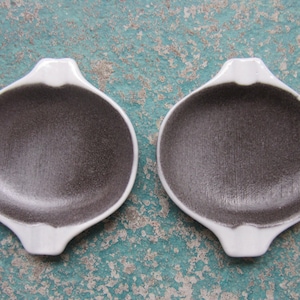 May include: Two small, white ceramic dishes with grey interiors. The dishes have a rounded shape with two small handles on either side.