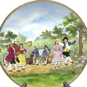 May include: A decorative plate featuring a pastoral scene with figures in period clothing dancing and playing instruments. The plate has a gold rim and is displayed on a black stand. The scene includes trees, a lake, and a small building.